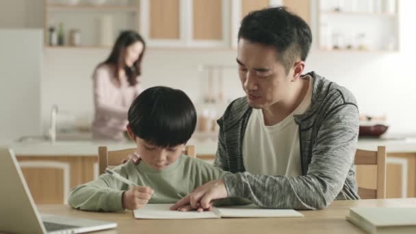 joven asiático padre ayudar hijo con tarea mientras madre preparación comida en cocina - Metraje, vídeo