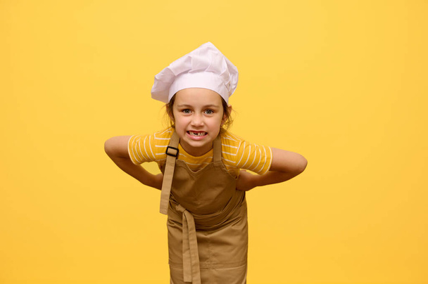 Rozpustilá holčička v kuchařově klobouku a zástěře, šklebící se, tvářící se, pózující s rukama kolem pasu, izolovaná na žlutém pozadí s reklamním prostorem. Koncept zábavy a vzdělávání pro děti. - Fotografie, Obrázek