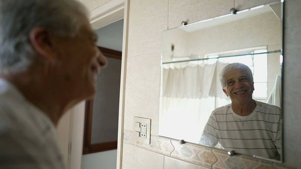 Счастливый старший мужчина смотрит на свое отражение ванной комнате улыбаясь, пожилой человек, начиная день с позитивным настроем - Фото, изображение