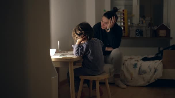 Откровенный ребенок, занимающийся домашним обучением ночью перед сном в пижаме, уставшая мать, наблюдающая за маленьким мальчиком, выполняют задание, чувствуя усталость - Кадры, видео