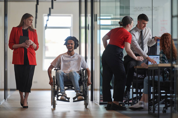 A group of young business people in a modern glass-walled office captures the essence of diversity and collaboration, while two colleagues, including an African American businessman in a wheelchair - Valokuva, kuva