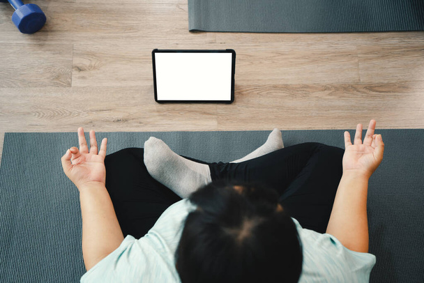 Cours de yoga à la maison avec tablette de technologie en ligne, vue du dessus de la jeune femme grosse assise sur le tapis de yoga et utilisant une tablette pendant la méditation pratiquant le yoga dans l'exercice de sukhasana au studio de yoga. - Photo, image