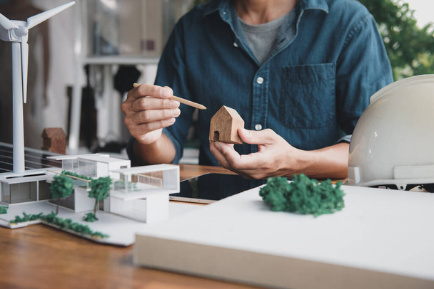 Architekt Designer halten kleine Holzhaus Symbol, während das Denken über Wohndesign kombiniert mit Wohnen mit natürlichen Energiekonzept, mit modernen Haus-und Windkraftanlage Modell, Sonnenkollektoren auf dem Schreibtisch. - Foto, Bild