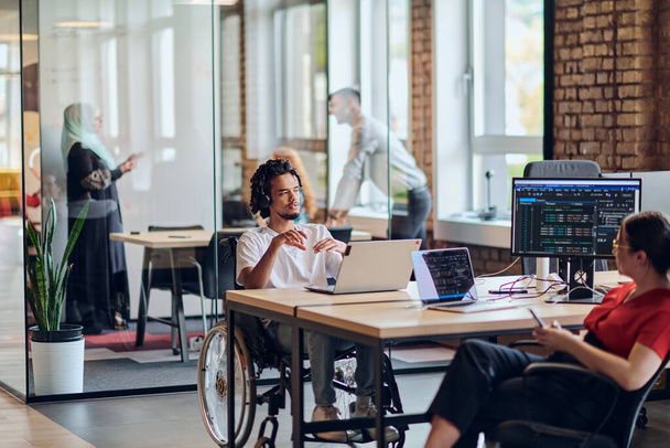 A young business group, including an African American businessman in a wheelchair, collaborates within a modern glass office, actively engaged around a computer and laptop, collectively solving - Foto, afbeelding