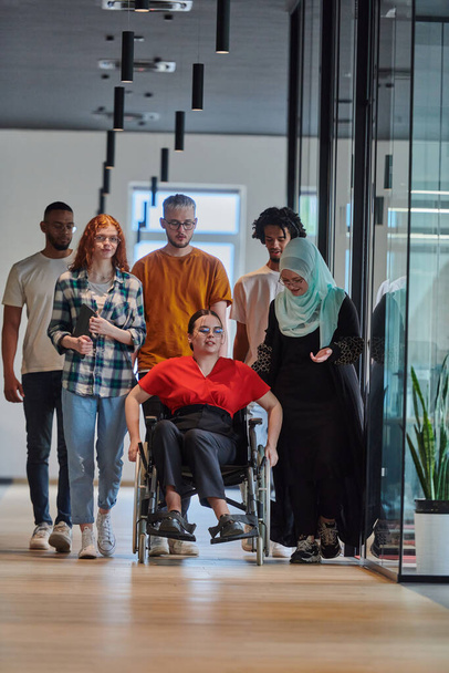 A diverse group of young business people walking a corridor in the glass-enclosed office of a modern startup, including a person in a wheelchair and a woman wearing a hijab, showing a dynamic mix of - Valokuva, kuva