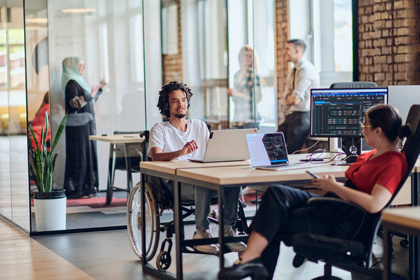 A young business group, including an African American businessman in a wheelchair, collaborates within a modern glass office, actively engaged around a computer and laptop, collectively solving - Foto, immagini