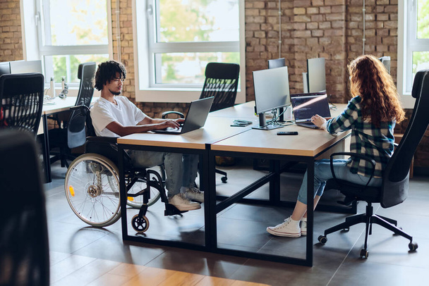 Business colleagues work within a modern startup coworking office, with one of them, an African America in a wheelchair, actively engaging with computers and laptops to collaboratively address various - Foto, Imagem