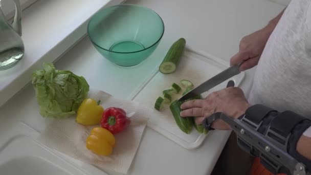 man with rigid elbow orthopedic holder with hinge, modern adjustable elbow orthosis, makes salad, peels, cuts vegetables with kitchen knife. - Footage, Video