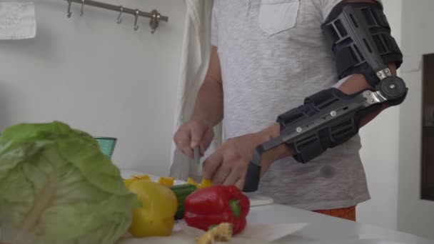 Mann mit starrem Ellbogenorthopädiehalter mit Scharnier, modern verstellbare Ellbogenorthese, macht Salat, schält, schneidet Gemüse mit Küchenmesser. - Filmmaterial, Video