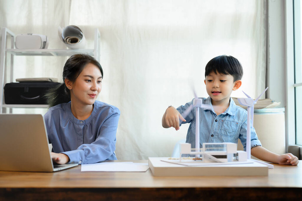 Little kid son aime jouer avec l'éolienne près de Young Asian mother is an energy and environmental engineer while working in home office, teaching and instilling ideas about natural energy. - Photo, image