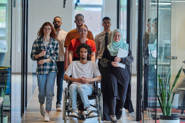 Modern bir girişimin cam kaplı ofisinde koridorda yürüyen bir grup genç iş adamı arasında tekerlekli sandalyedeki bir kişi ve tesettürlü bir kadın da var. - Fotoğraf, Görsel