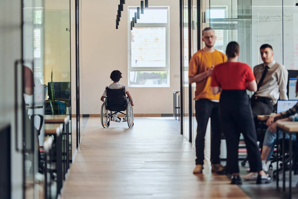 Monipuolinen joukko kollegoita keskustelee liiketoiminnan haasteista modernissa coworking startup-keskuksessa, kun taas taustalla heidän pyörätuolilla kulkeva kollegansa symboloi - Valokuva, kuva