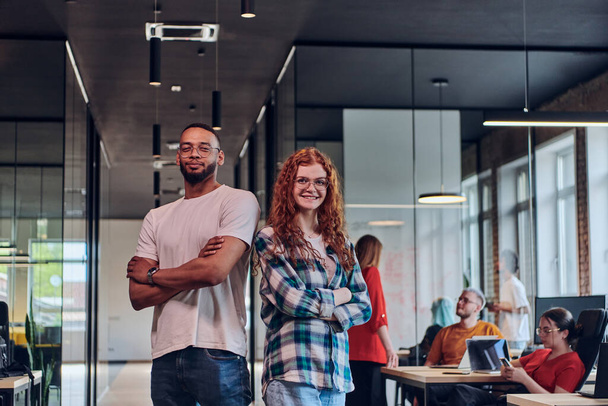 A young African American businessman and a modern businesswoman with orange hair stand side by side, arms crossed, exuding confidence and unity in a contemporary office setting, epitomizing dynamic - 写真・画像