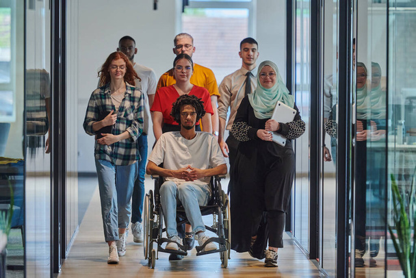 A diverse group of young business people walking a corridor in the glass-enclosed office of a modern startup, including a person in a wheelchair and a woman wearing a hijab, showing a dynamic mix of - Fotoğraf, Görsel