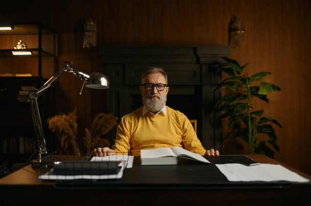 Портрет серьезного задумчивого пожилого человека на пенсии, работающего в домашнем офисе вечером. Пожилой мужчина, сидящий за столом и читающий книги в библиотеке - Фото, изображение