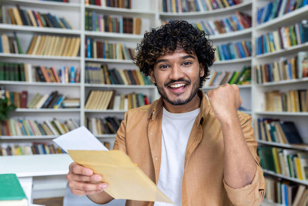 Portrait of young successful student, man received letter of notification of good results of interview exam, hispanic man joyfully looking at camera inside library, holding mail envelope. - Fotó, kép