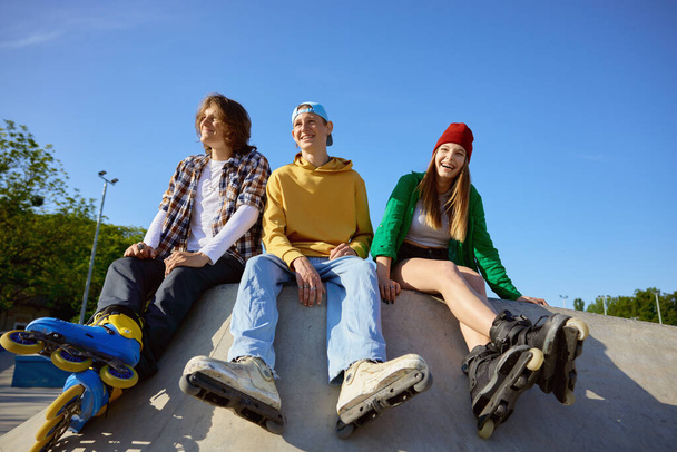 Joyful rollerskaters team of teenage people sitting on ramp in skate park. Extreme sports training and processional equipment for teens - 写真・画像