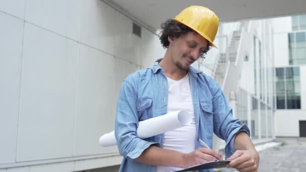 architekt muž konstrukce inženýr pracovník nebo inspektor stojí venku před moderní budovy s ochrannou přilbou na hlavě a dokumenty v rukou rozhlíží - Záběry, video