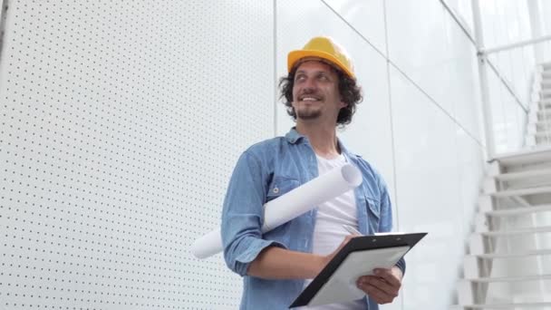 építész férfi építőmérnök munkás vagy ellenőr áll a szabadban, a modern épület előtt védősisakkal a fején, és dokumentumok a kezében körülnéz - Felvétel, videó