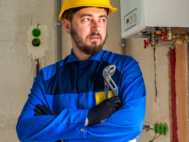 Retrato de trabajador o ingeniero en ropa de trabajo azul y casco de seguridad amarillo con espacio para texto. Un hombre en un sitio de construcción sostiene una llave de tubo ajustable en sus manos y mira hacia otro lado. - Foto, imagen