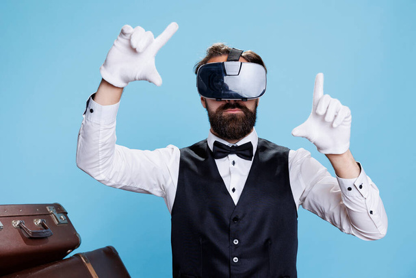 Portero moderno con auriculares vr contra fondo azul, disfrutando de la experiencia de visión interactiva 3d. Trabajador profesional de hotel botones divirtiéndose con gafas de realidad virtual. - Foto, imagen