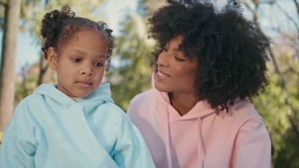 Boldog anya csók lánya ül a gyönyörű napos természet közelről. Gondtalan afro-amerikai gyermek élvezi a családi pikniket a kedves anyukájával. Mosolygó nő csodálta gyerek kifejező szeretet gyengédség. - Felvétel, videó