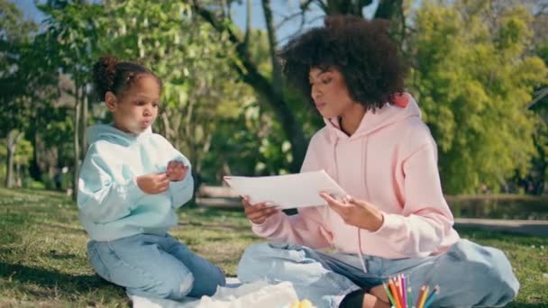 Woman daughter eating on picnic sitting sunny green lawn close up. Carefree african american mother giving paper for drawing to cute chewing girl outdoors. Relaxed happy family enjoy weekend on nature - Footage, Video