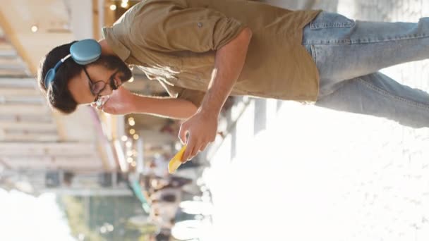 Feliz hombre indio relajado y alegre en la elección de auriculares inalámbricos, escuchando música disco energética favorita en el baile de teléfonos inteligentes. Guy paseo turístico pasa por la calle urbana sol de la ciudad. Vertical - Imágenes, Vídeo