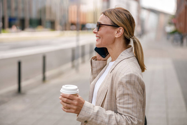 Усміхнена жінка офіс працівник розмовляє телефоном на сучасному будівельному фоні і п'є каву - Фото, зображення