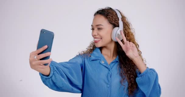 Woman, peace or headphones for selfie in studio, live streaming or video call on white background. Profile picture, happy gen z model and v sign with tongue out, blowing kiss or emoji on social media. - Footage, Video