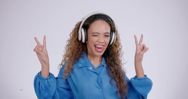 Woman, funny face and peace sign with music in studio with a smile with v hand gesture. Grey background, comic and female person with portrait and excited with headphones and happy emoji hands. - Footage, Video