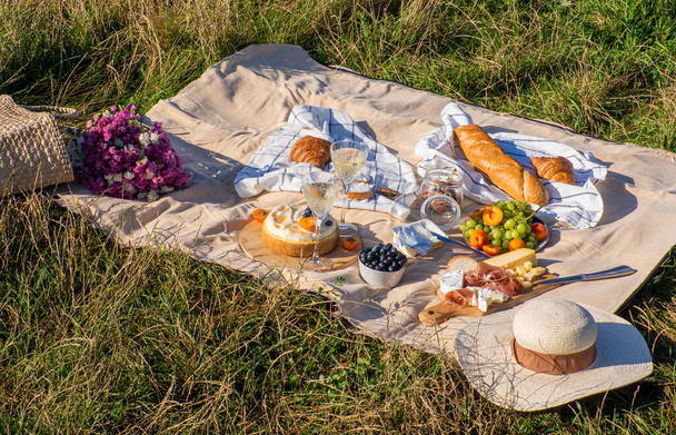 Beige blanket with fruits and pastries, cookies, berries, cheese, straw hat, baguette and bag with pink flowers. Concept of having picnic in a city park during summer holidays or weekends. Copy space. - Photo, Image