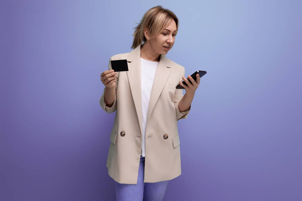 Blonde junge Frau in Jacke bezahlt Einkäufe mit Telefon und Karte vor Studiohintergrund. - Foto, Bild