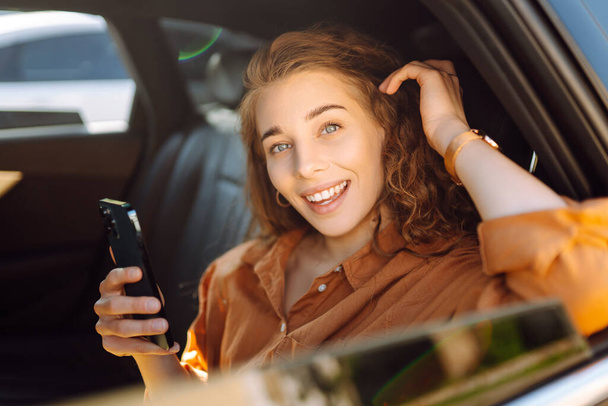 Cheerful woman with curly hair and stylish clothes sits in the back seat of a car, uses a mobile phone. The concept of business, technology, travel, online communication. - Fotografie, Obrázek