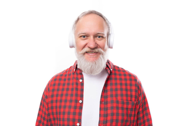 Un pensionista canoso de 60 años con barba y bigote en camisa escucha música en auriculares sin cable. - Foto, imagen