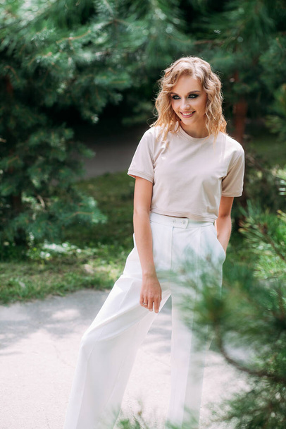 Стройная улыбающаяся молодая девушка с модным макияжем и стильной прической, в белых брюках и бежевом топе, стоит на фоне парка и игриво смотрит в камеру. - Фото, изображение
