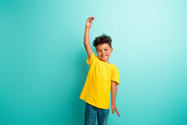 Photo of cheerful schoolboy with brown hair dressed yellow t-shirt raising arm up measure height isolated on turquoise color background. - Foto, Imagen