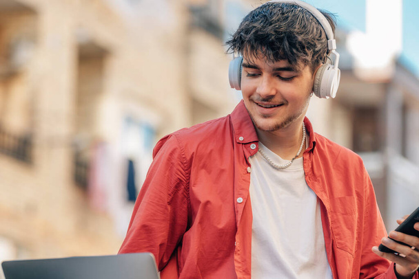 fiatal férfi okostelefonnal vagy mobiltelefonnal, laptoppal és fejhallgatóval a szabadban a városban - Fotó, kép