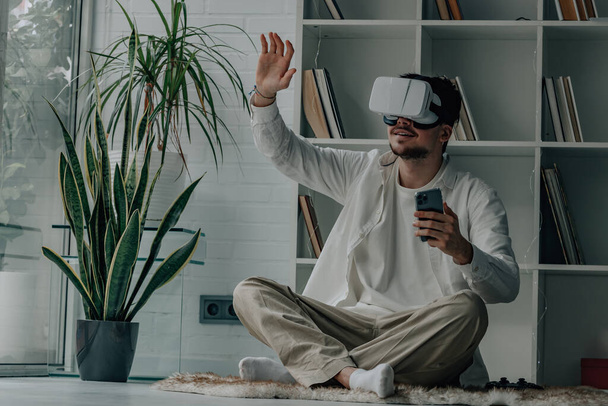 Młody człowiek w domu w okularach wirtualnej rzeczywistości i telefonie komórkowym - Zdjęcie, obraz