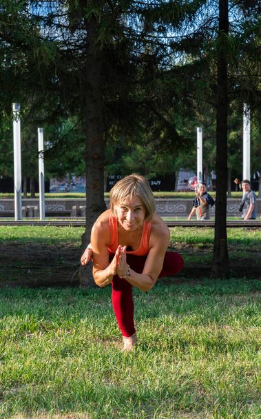 Şehir caddesinde çimlerin üzerinde yoga yapan 59 yaşındaki olgun bir kadının portresi. Yetişkinlerin aktif spor yaşam tarzı.  - Fotoğraf, Görsel