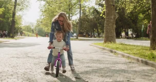 Madre joven enseña a su hijo pequeño a montar bicicleta de equilibrio en el parque de la ciudad. Rubia padre apoya niño montar en bicicleta carrera en el pavimento soleado en el parque - Imágenes, Vídeo
