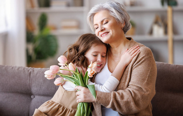 花の花束を持つ幸せなシニア祖母は優しく彼のかわいい孫娘を抱擁し、お祝いと祖父母の日に休日を取得しながら彼女からの贈り物を受け取ります - 写真・画像