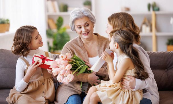 Fijne Internationale Vrouwendag. Glimlachende multi generatie familie dochter en kleindochter geven bloemen en cadeau aan grootmoeder vrolijk vieren de voorjaarsvakantie Moederdag of grootouders dag thuis - Foto, afbeelding