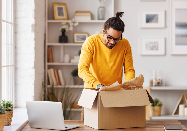 Hombre afroamericano en ropa casual con gafas sonriendo y desempacando pedido entregado de caja de cartón mientras está en la mesa en la oficina en casa - Foto, Imagen