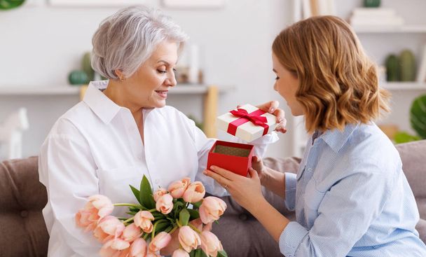 Positiv elegante ältere Frau mit einem Bündel frischer Tulpen lächelt und nimmt ein Geschenk einer kleinen Tochter während einer Muttertagsfeier entgegen - Foto, Bild