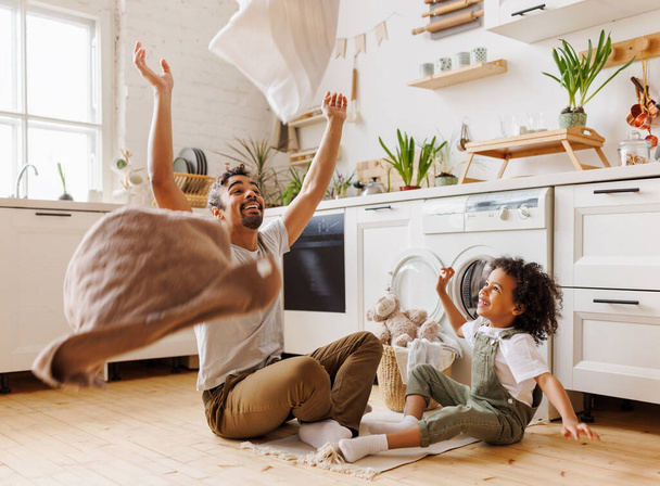 Happy african american dad and son smiling and tossing towels in air while sitting on floor near washing machine during household routine in kitchen at home - Photo, Image