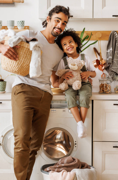 Alegre papá negro con cesta de lavandería mirando a la cámara con sonrisa y abrazando al niño optimista mientras hace las tareas domésticas cerca de la lavadora en la cocina ligera en casa - Foto, imagen