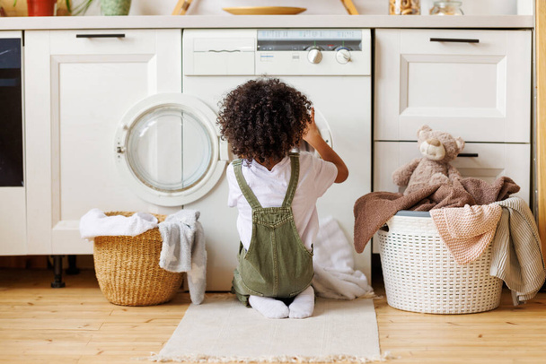 Πίσω όψη του μαύρου αγοριού με σγουρά μαλλιά γονατιστό στο πάτωμα και βάζοντας ρούχα στο πλυντήριο ενώ κάνει πλυντήριο στην κουζίνα στο σπίτι - Φωτογραφία, εικόνα
