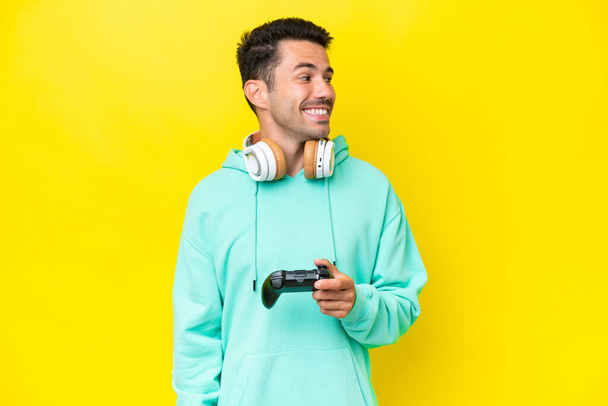 Jeune homme beau jouant avec une manette de jeu vidéo sur un mur isolé regardant sur le côté et souriant - Photo, image