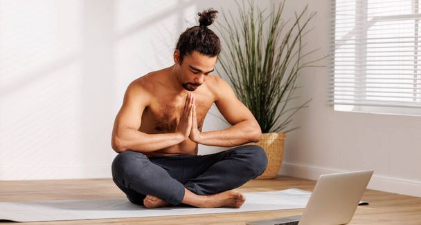  Jonge vrolijke Afrikaanse Amerikaanse man met naakte romp zittend in lotus poseren voor laptop en dank leraar in de woonkamer thuis, met yoga klasse online. Het kijken naar Yoga Tutorials Online - Foto, afbeelding